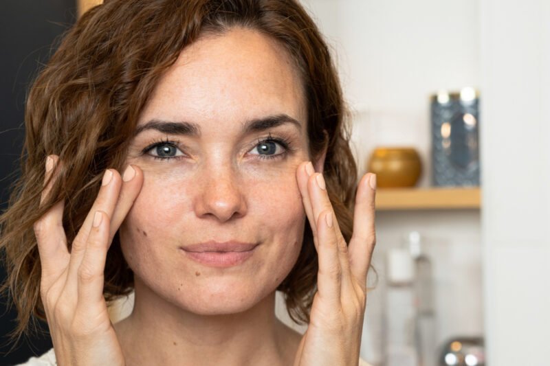 Woman touching skin under eyes looking at her face in the mirror