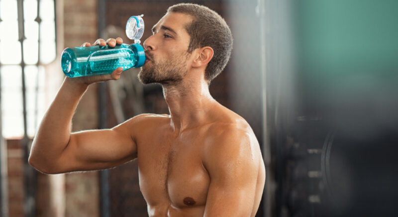 A young, fit shirtless man drinking water at the gym